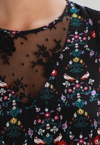 Vestido negro con un estampado de flores y pájaros de colores, que presenta un escote de encaje negro semitransparente con bordado floral.