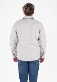 Camicia button-up grigia chiara con maniche lunghe, colletto casual e cuciture accentate nere. Indossata con jeans blu, che mostrano una vestibilità rilassata.