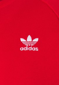 Röd stickad tyg med en vit Adidas trefoil-logotyp och text i gemener broderad i mitten.