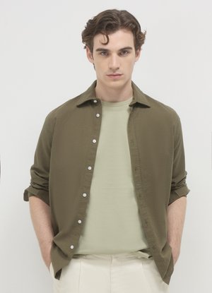 Chemise boutonnée vert olive portée sur un t-shirt à col rond vert clair. La chemise présente des boutons blancs et des manches retroussées, portée avec un pantalon beige.