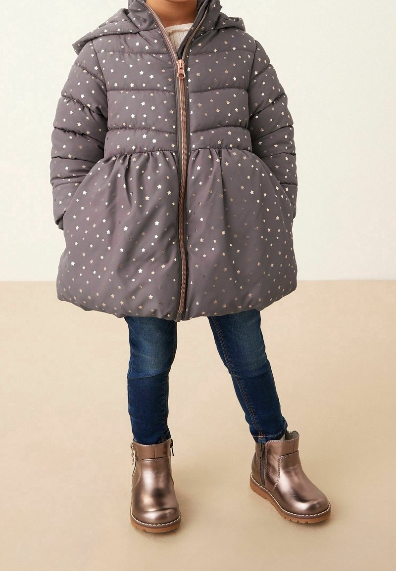 Enfant portant une doudoune grise à motif étoilé, un jean bleu foncé et des bottines métalliques bronze, debout devant un fond uni.