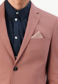 Ανοιχτό ροζ σακάκι με λεία υφή, με ένα μαύρο κουμπί και τσέπη στο στήθος που κρατάει ένα ανοιχτό μπεζ μαντήλι, φορεμένο πάνω από ένα ναυτικό πουκάμισο.