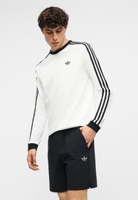 Young man wearing white long-sleeve Adidas sweatshirt with black stripes and black Adidas shorts, standing with one hand in pocket.