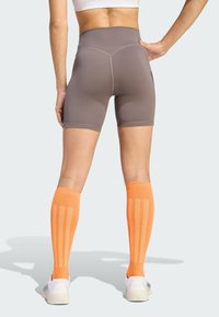 Sportke hlače smeđe boje s visokim strukom uparene s narančastim čarapama do koljena s okomitim prugama i bijelim sportskim tenisicama. Glatka tekstura tkanine.