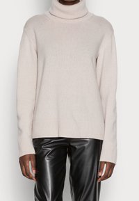 Ljusbeige polotröja i mjukt stickat material. Har ribbad textur, nedsänkta axlar och en rak fåll. Kombinerad med svarta skinnbyxor.