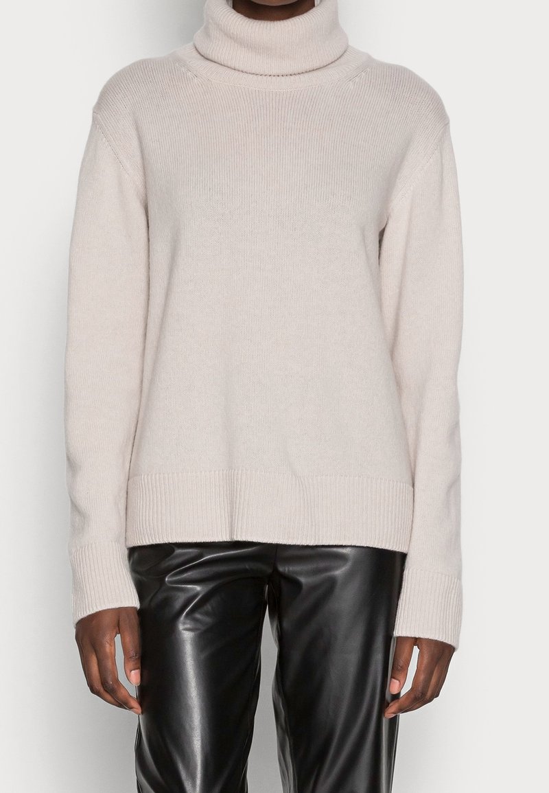 Ljusbeige polotröja i mjukt stickat material. Har ribbad textur, nedsänkta axlar och en rak fåll. Kombinerad med svarta skinnbyxor.