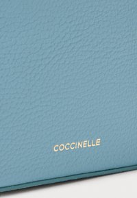 Suprafață de piele texturată albastră cu un logo „Coccinelle” embosat în aur în partea de jos. Materialul pare moale și delicat.