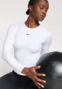 Bijela atletska majica dugih rukava s okruglim izrezom i uskim krojem, s malim crnim Nike logom. Drži se crna medicinska lopta.
