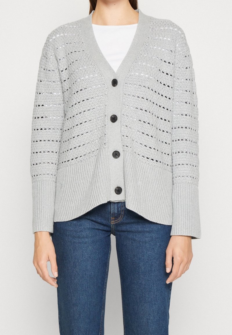 Femme portant un cardigan boutonné en maille ouverte gris clair par-dessus un haut blanc avec un jean bleu, cadrée au niveau du cou et mi-cuisses.
