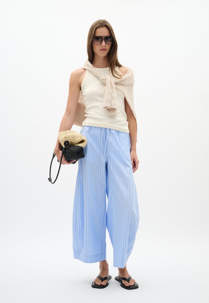 Femme portant un haut beige sans manches avec un pull assorti sur les épaules, un pantalon large rayé bleu clair, des sandales noires et de grandes lunettes de soleil, tenant des sacs tissés.