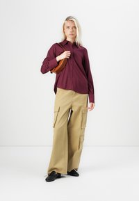 Maroon button-up shirt, loose beige cargo pants with pockets, and black shoes. Model carries a small brown shoulder bag.