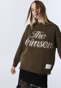 Brauner gestrickter Pullover mit dem Schriftzug "The Crimson" in mehrfarbigem Stick. Ausfransungen am Saum, kombiniert mit einem Spitzenrock.