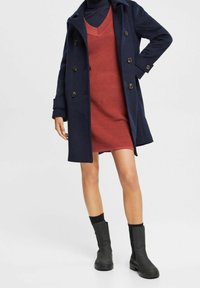 Cappotto navy con grandi bottoni, indossato sopra un abito in maglia color ruggine. Stivali neri alla caviglia completano l'outfit, mostrando un look a strati.