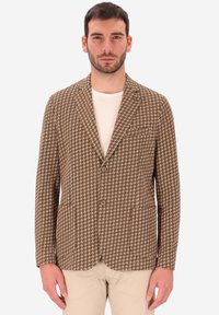 Uomo che indossa un blazer a scacchi marrone e bianco sopra una camicia bianca con pantaloni beige, in piedi davanti a uno sfondo chiaro e uniforme.