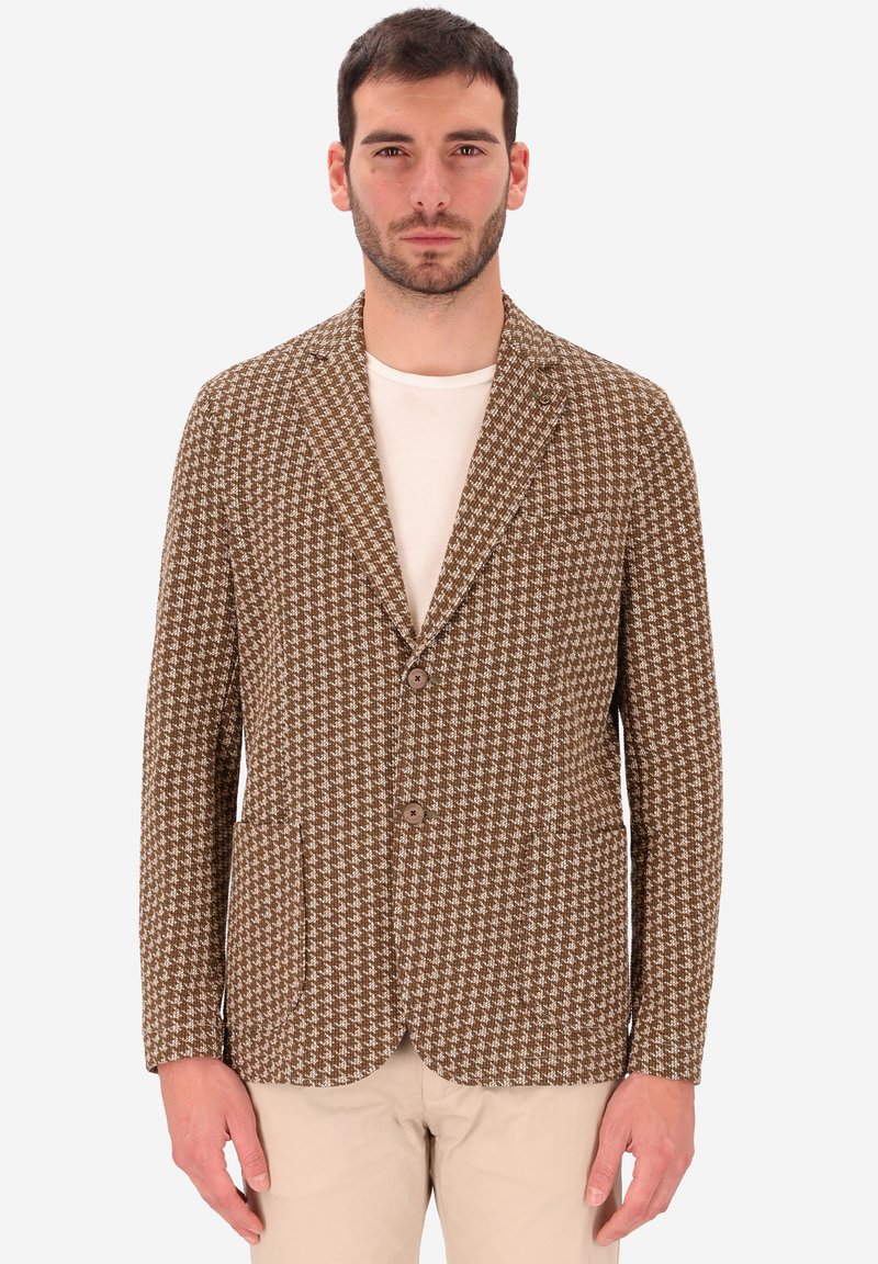 Uomo che indossa un blazer a scacchi marrone e bianco sopra una camicia bianca con pantaloni beige, in piedi davanti a uno sfondo chiaro e uniforme.