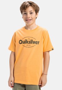 Oranssi puuvillapaita lyhythihaisilla hihoilla, jossa on keskiössä musta graafinen printti, jossa lukee "Quiksilver" ja pieni logo hihassa.