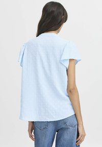 Blusa azzurro chiaro con motivo a pois texture, maniche corte a sbuffo e orlo arrotondato. Tessuto morbido, vestibilità casual e semplice dettaglio del colletto.