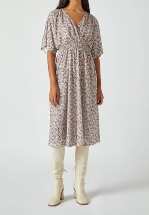 Woman wearing a floral midi dress with elbow-length sleeves and cream knee-high heeled boots, standing against a plain background.