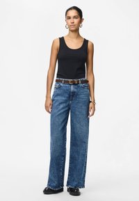 Γυναίκα που στέκεται, φοράει μαύρο τοπ χωρίς μανίκια, ψηλόμεσα μπλε φαρδιά τζιν με καφέ ζώνη, μαύρα παπούτσια και χρυσούς κρίκους σκουλαρίκια.