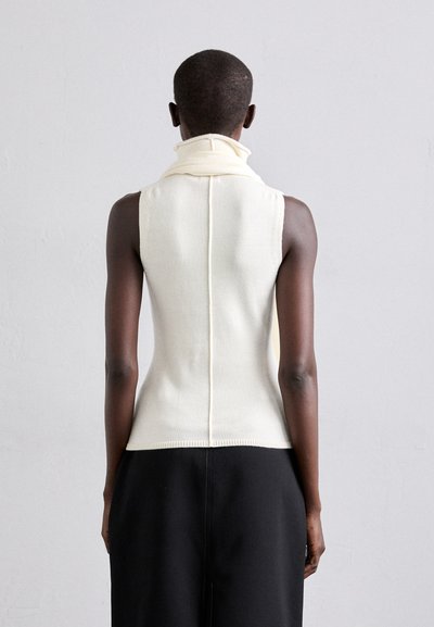 Top tricot sans manches crème avec col montant, présentant une coupe ajustée et un ourlet côtelé, associé à un pantalon noir ample. Texture lisse et design minimaliste.