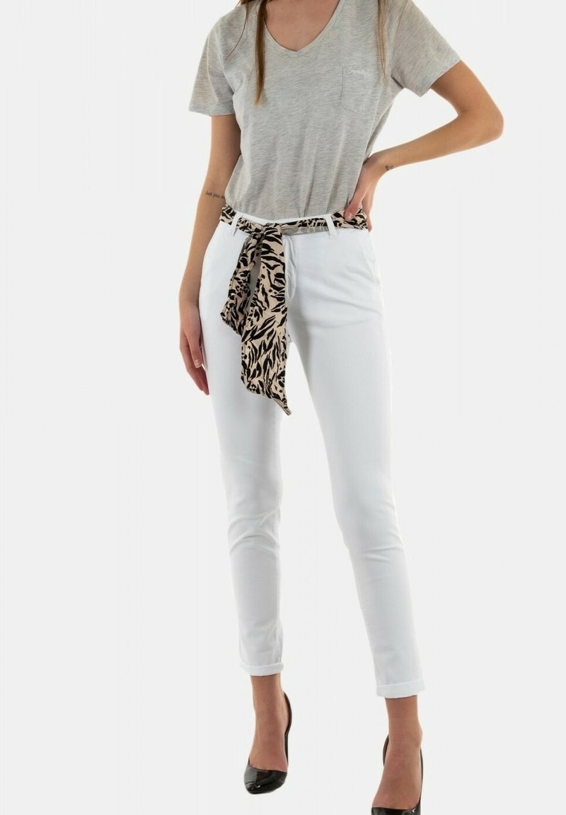 Femme portant un t-shirt beige à col en V, un pantalon blanc avec une ceinture imprimé léopard nouée et pendante à l'avant, et des chaussures à talons noires.