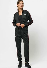 Fekete, sima anyagból készült atlétikai melegítő, piros oldalsávokkal. Cipzáros felső és elasztikus derékpántú nadrág, a mellrészen logóval.