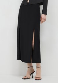 Jupe midi noire avec fentes latérales, tissu lisse, design minimaliste, taille haute, associée à des talons aiguilles noirs à lanières.