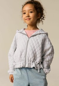 Haut de sweatshirt zippé gris clair matelassé avec capuche, doté d'une taille et de poignets élastiques, porté sur un top rose et un short en denim bleu clair.