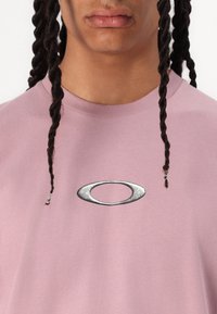 T-shirt rosa claro com um logotipo metálico redondo centrado no peito. Feito de tecido macio, com decote redondo e mangas curtas.