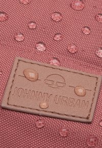 Rózsaszín textúrájú szövet vízcseppekkel és egy világosbarna bőr folttal, amelyen egyszerű betűtípussal a "JOHNNY URBAN" felirat található.