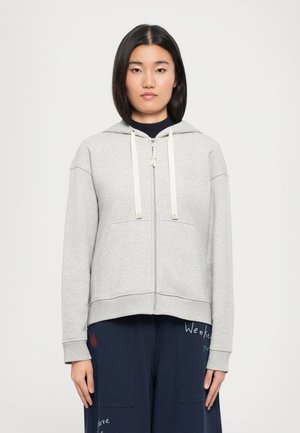 Hellgraue Zip-Hoodie mit Kapuze, ausgestattet mit weißen Kordeln, Fronttaschen und gerippten Bündchen. Über einer dunkelblauen Hose mit Stickerei getragen.