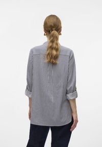 Kvinde med langt bølget hår samlet i en hestehale, iført en vertikalt stribet skjorte med optrukne ærmer og mørke bukser, set bagfra.