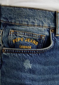 Siniset farkut, joissa on haalistunut pinta, varustettuna ommellulla taskulla, jossa lukee keltaisilla kirjaimilla "PEPE JEANS LONDON".