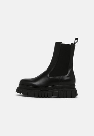 Botte Chelsea en cuir noir avec semelle carrée crantée, panneaux élastiques latéraux et languette de traction à l'arrière, vue de profil sur fond blanc.