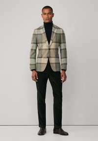 Blazer de tweed con un patrón a cuadros en azul, amarillo y marrón, combinado con un cuello de tortuga negro y pantalones en verde oscuro. Zapatos marrones completan el conjunto.