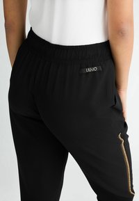 Czarne spodnie z elastyczną talią o dopasowanym kroju, z ozdobnym złotym suwakiem po boku i oznakowaną metką z tyłu.