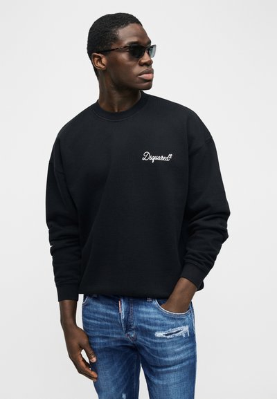 Άνδρας με μαύρο φούτερ Dsquared2, μπλε τζιν με φθορές και μαύρα γυαλιά ηλίου, στέκεται με το ένα χέρι στην τσέπη.