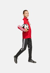 Rote Zip-Jacke mit weißen Akzenten und drei Streifen an den Ärmeln, kombiniert mit schwarzen Hosen mit weißen Streifen und schwarzen Fußballschuhen, während er einen Fußball hält.