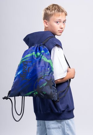 Boy wearing a blue sleeveless hoodie and carrying a blue and green drawstring backpack with a soccer ball design, looking over his shoulder.