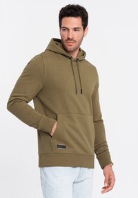 Sudadera verde oliva con un bolsillo frontal, capucha ajustable con cordón, puños de canalé y un pequeño detalle de etiqueta negra. Textura suave, corte informal.