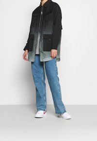 Giacca ombre nera e grigia con due tasche frontali e chiusura a zip, indossata sopra una maglietta grigia e abbinata a jeans in denim blu e sneaker bianche.