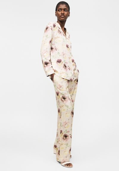 Γυναίκα με μπλούζα μακρυμάνικη κρεμ χρώματος με λουλούδια και ταιριαστό φαρδύ παντελόνι, στέκεται με τα χέρια στις τσέπες, στραμμένη ελαφρώς δεξιά.