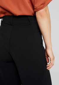 Pantalon noir sur-mesure avec une texture lisse, taille haute et passants de ceinture, vu de dos avec un haut orange à manches courtes partiellement visible.