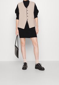 Veste sans manches beige avec des boutons superposée sur une robe en maille noire, assortie à des chaussures noires et des chaussettes claires. Sac à main noir tenu à la main.