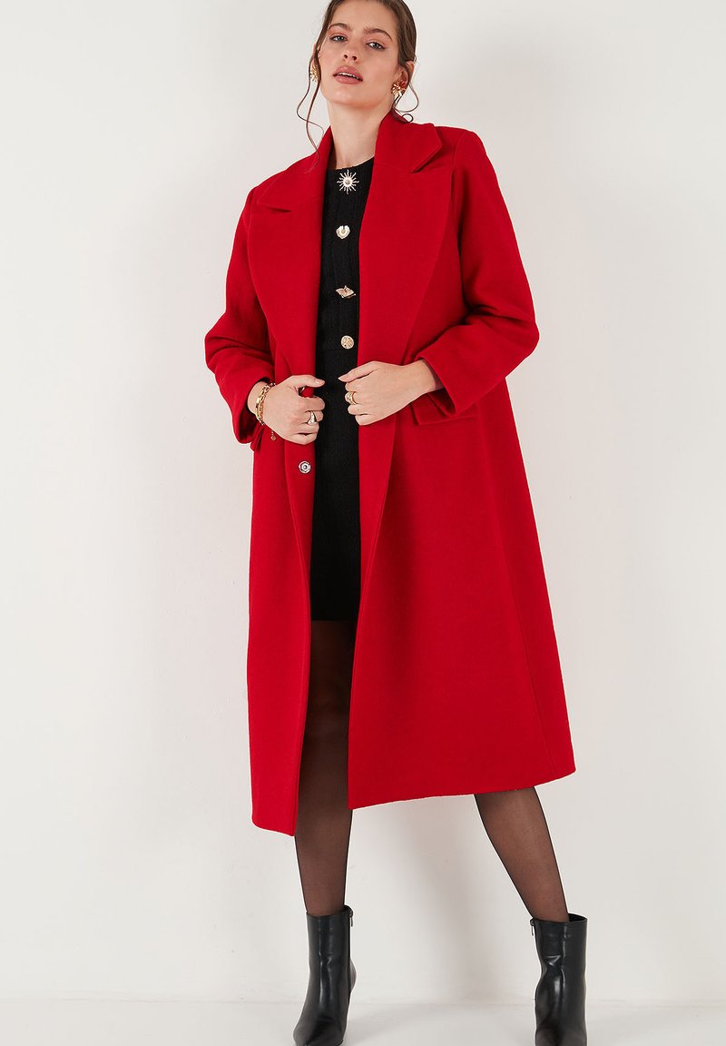Red wool coat with large notched lapels and long sleeves. Features side pockets and a tailored fit. Worn over a black dress and ankle boots.