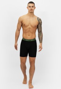 Schwarze Boxershorts mit einem grünen Bund, der ein Logo zeigt. Das Material scheint glatt und dehnbar zu sein, konzipiert für Komfort.