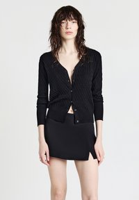 Cardigan nero con un motivo tessuto, dotato di scollatura a V, maniche lunghe e chiusure a bottoni, abbinato a una minigonna nera.