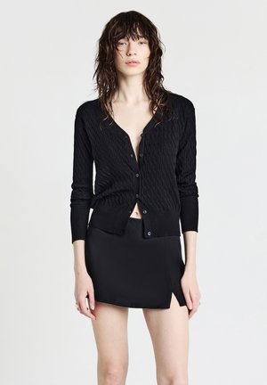 Schwarze Strickjacke mit strukturiertem Muster, V-Ausschnitt, langen Ärmeln und Knopfverschluss, kombiniert mit einem schwarzen Minirock.