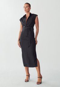 Vestido negro de camisa con mangas en Cap, frente abotonado, cintura ceñida con cinturón, largo midi, abertura lateral, textura suave, combinado con sandalias negras de tacón.