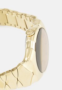 Montre dorée avec un boîtier rond, des maillons texturés sur le bracelet et un cadran en verre foncé avec une couronne latérale pour les réglages.